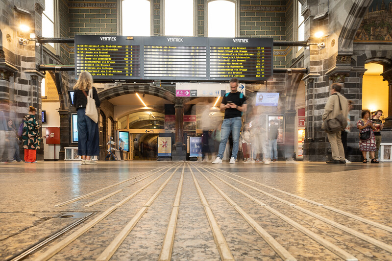 Enkele mensen lopen in de inkomhal van station Gent Sint-Pieters, waar ook het dienstregelingsbord te zien is.