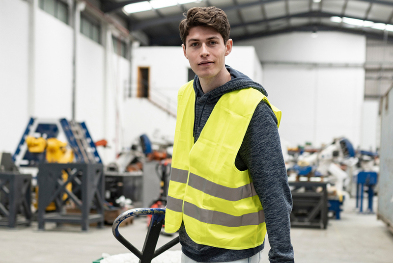Een jonge interimmer met een geel fluohesje aan, trekt een transpallet voort in een fabriek.