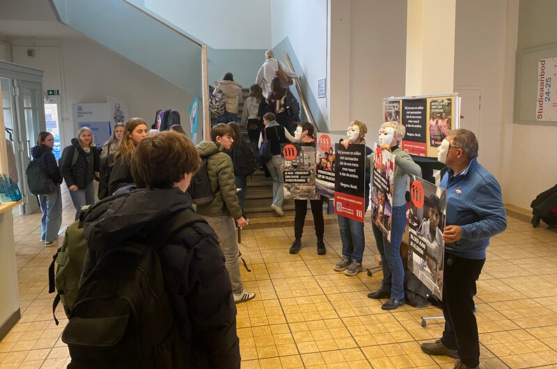 Gemaskerde vrijwilligers van 11.11.11 staan met pamfletten over menselijkheid in de trappenhal van een school.