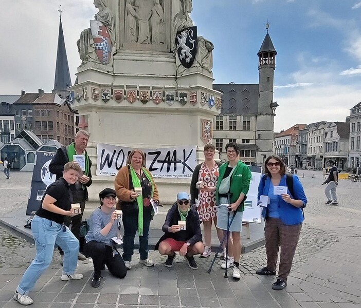 Acht personen poseren met een speldoosje in de hand voor een spandoek van de Woonzaak aan het standbeeld van Jacob Van Artevelde op de Vrijdagsmarkt in Gent.