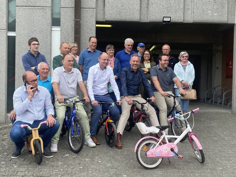 Een groep van 16 mensen staan gegroepeerd met op twee rijen. De voorste mensen zitten op een kinderfietsje.