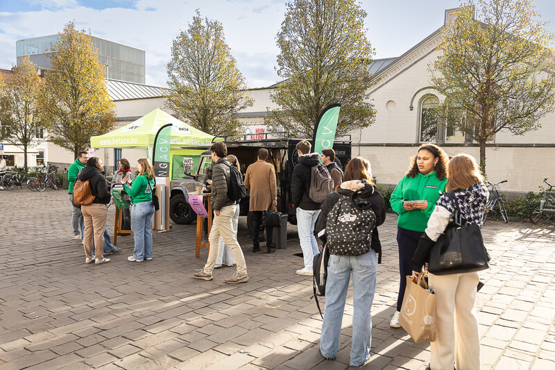 Op het Kramersplein in Gent staan vijf bomen in herfstkleuren en een oud wit gebouw in de achtergrond van deze foto. Het is zonnig weer. Op de voorgrond zien we een ACV-tentje en 3 Jong ACV ‘ers die met jongeren in gesprek gaan over hun inkomen.