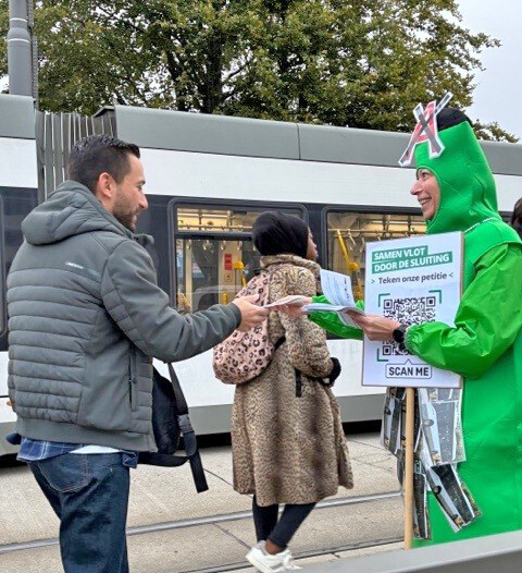 Aan een tramhalte deelt Carolien Vereeken van ACV, verkleed in een groene flessenhals, een petitiefolder uit aan tramreizigers.