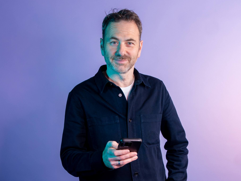 Tim Verheyden journalist van VRT staat op paarse achtergrond met telefoon in de hand. 