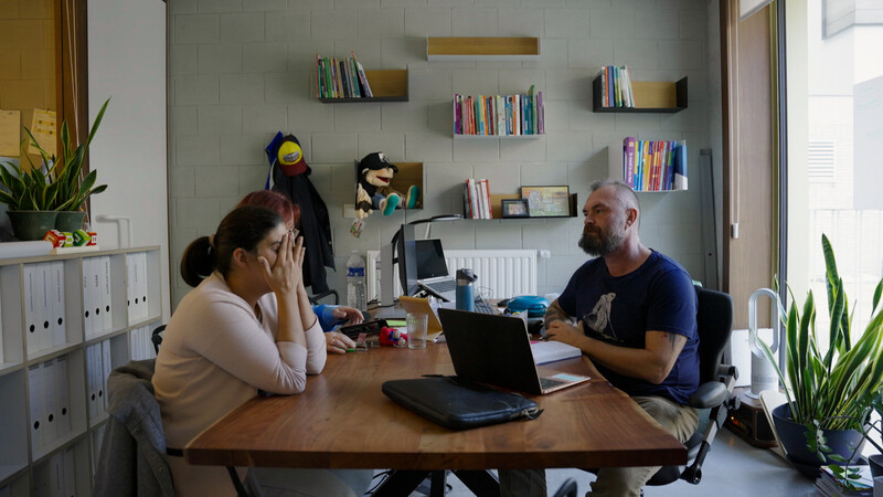 Vrouw en man zitten aan bureau. Vrouw houdt handen tegen gezicht, terwijl man luistert