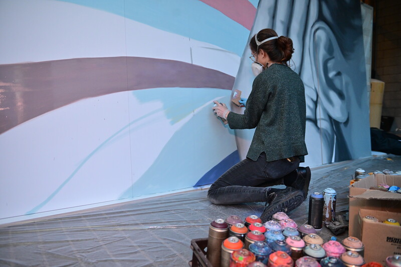 Een grafitti-kunstenares spuit verf en maakt een mural over het thema 'We houden je in het oor'.