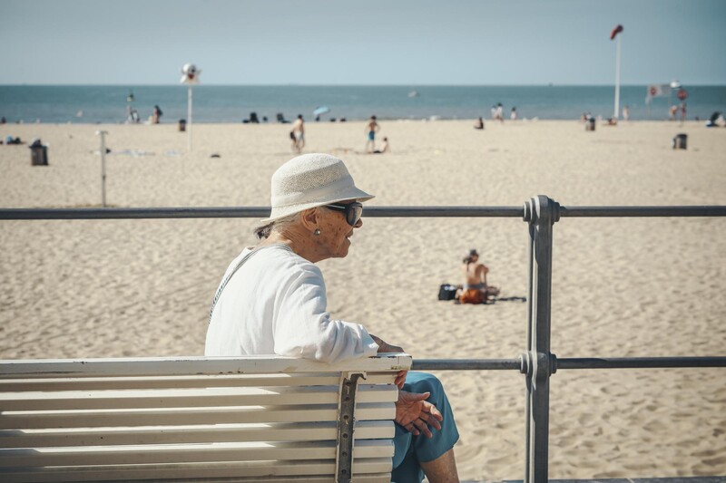 Oudere dame met hoed en zonnebril op bankje aan het strand
