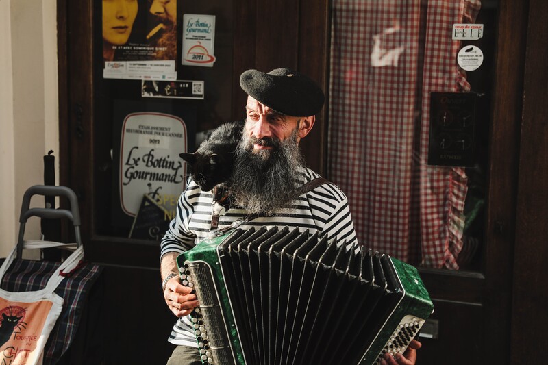 Man met beret en streepjestrui en zwarte kat op zijn schouder speelt accordeon in een Frans café