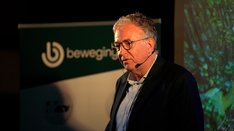 Luc Van Gorp speecht op podium voor banner van beweging.net