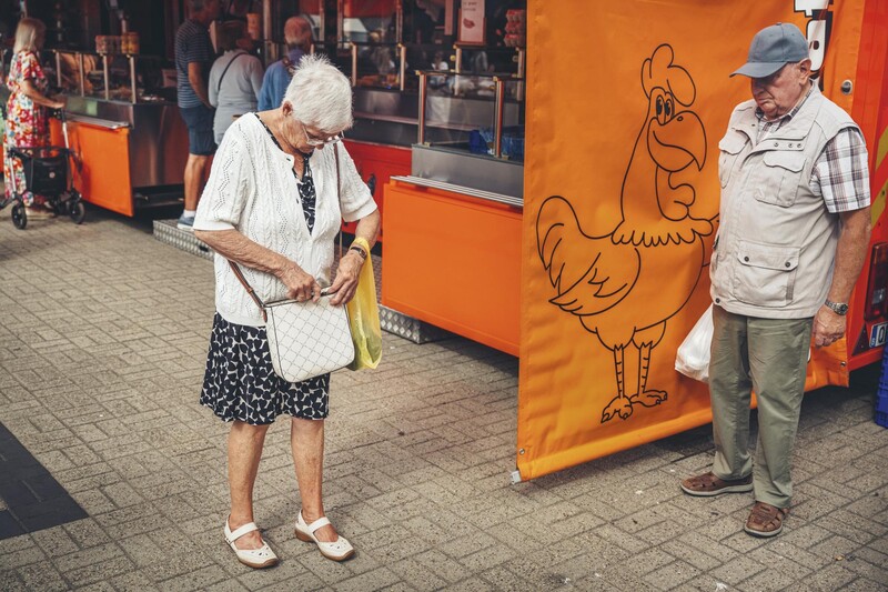 oudere vrouw kijkt in handtas op markt terwijl oudere man naar haar kijkt