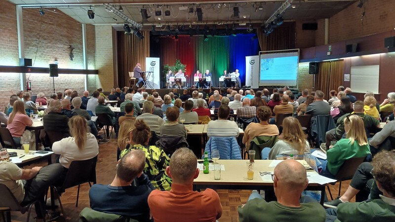 Een zaal met zittende mensen aan tafels, luisteren naar een politiek debat dat gaande is op het podium.