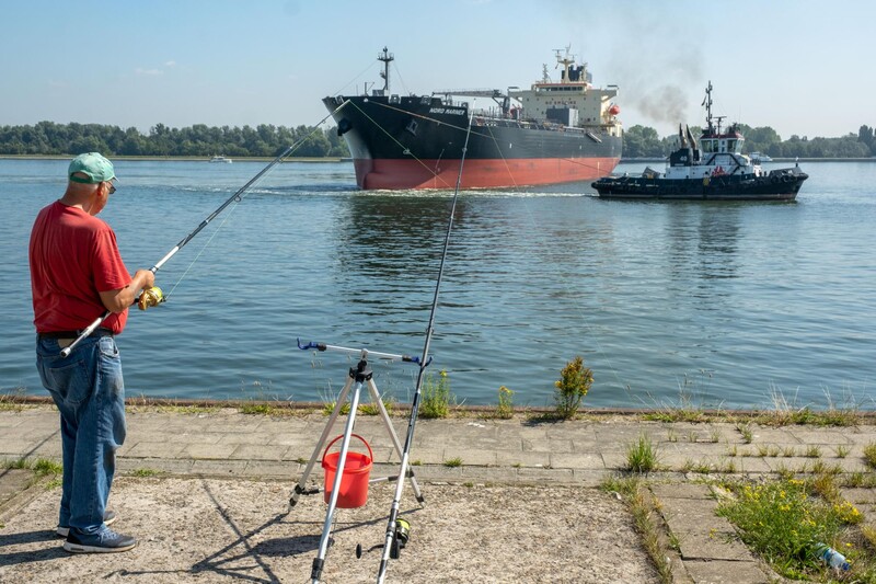 Groot schip met sleepboot en in voorgrond een visser op de kade