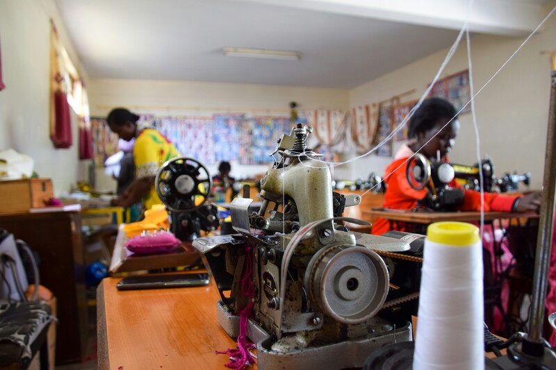 close-up van een naaitoestel met in de achtergrond een Congolees naai-atelier met enkele werkende vrouwen