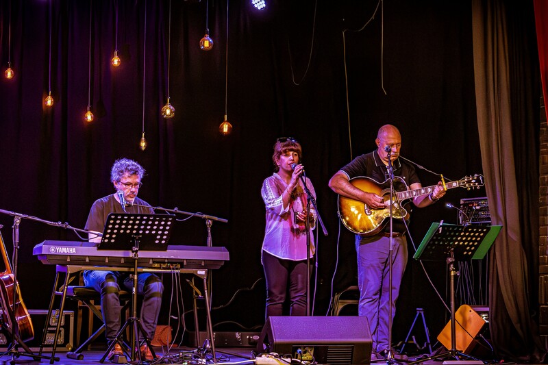 Donker podium met sfeerlicht en drie muzikanten op synthesizer, zang en gitaar.